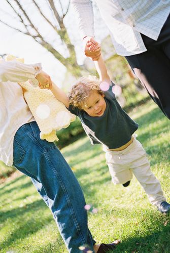 Two parents swing a curly-haired toddler by the hands in a sunny green park.
