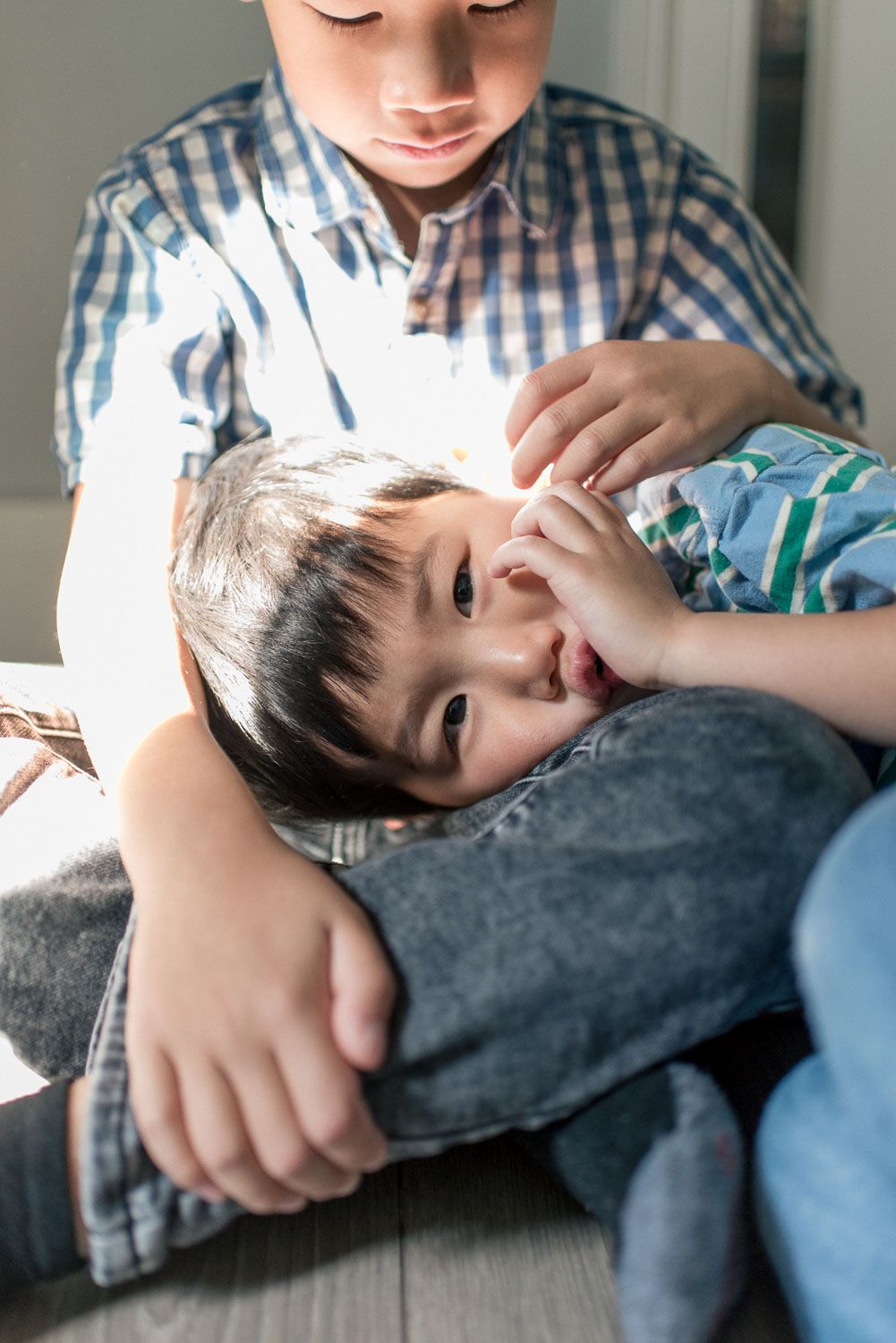 Two Asian boys sitting on floor, younger child resting head on older sibling's lap in a tender moment.