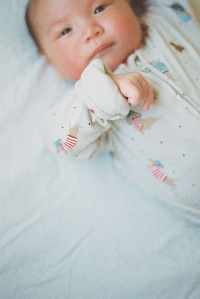 A newborn baby in a patterned onesie lies on a light blue blanket, raising a tiny fist toward the camera.