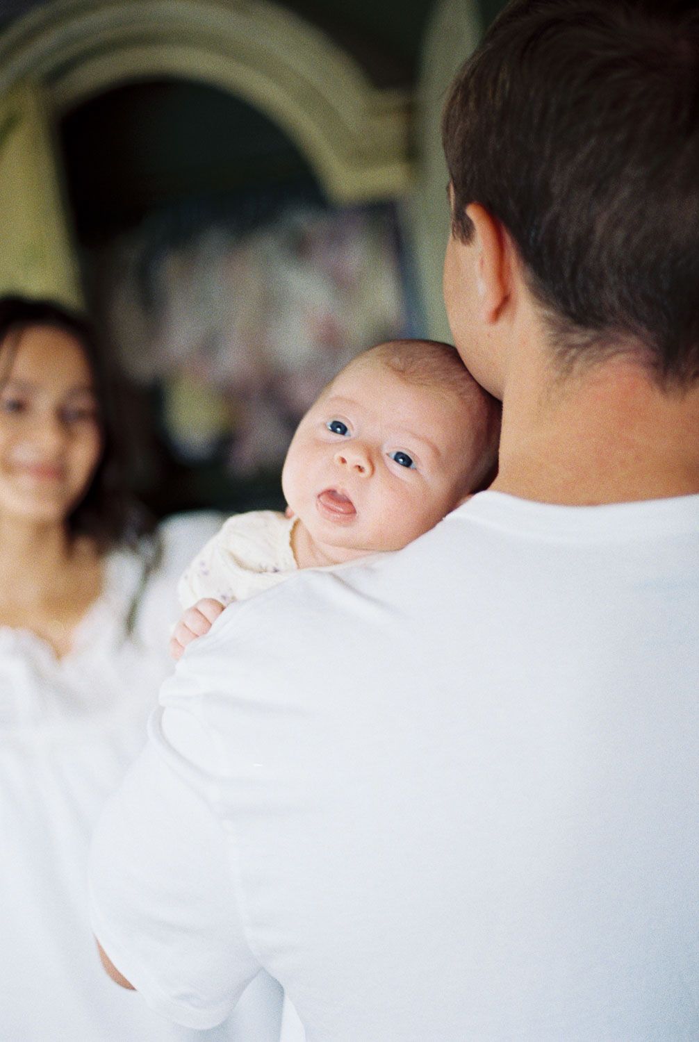 A father holds his newborn baby while a smiling mother looks on during an intimate family portrait session.