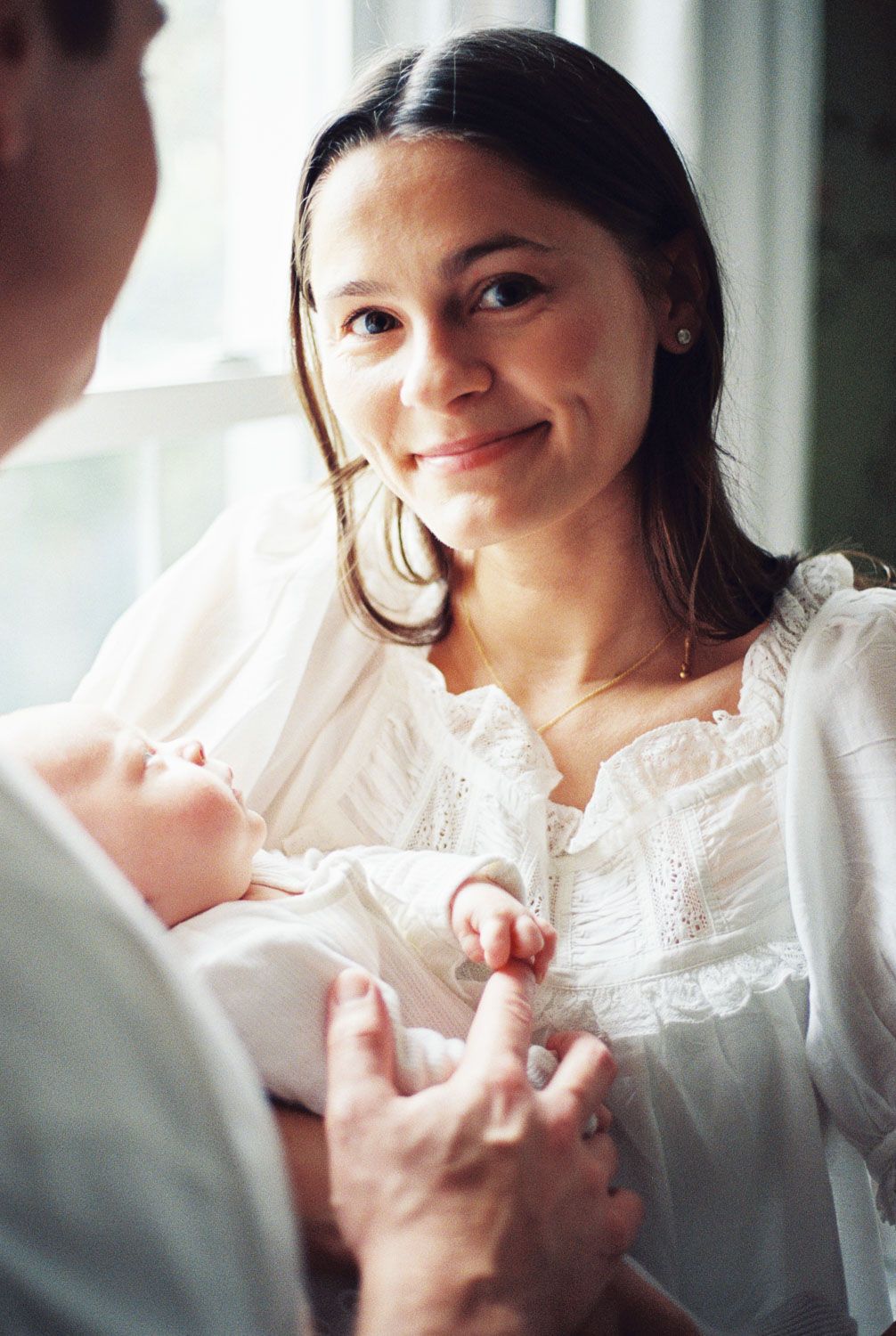 Smiling mother in white dress holds newborn baby while interacting with another person near a bright window.