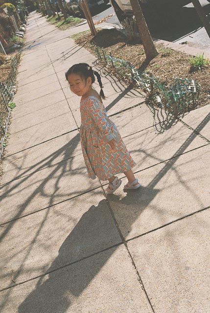 Young Asian girl in floral dress walking on sunny sidewalk, casting long shadow on concrete pavement.