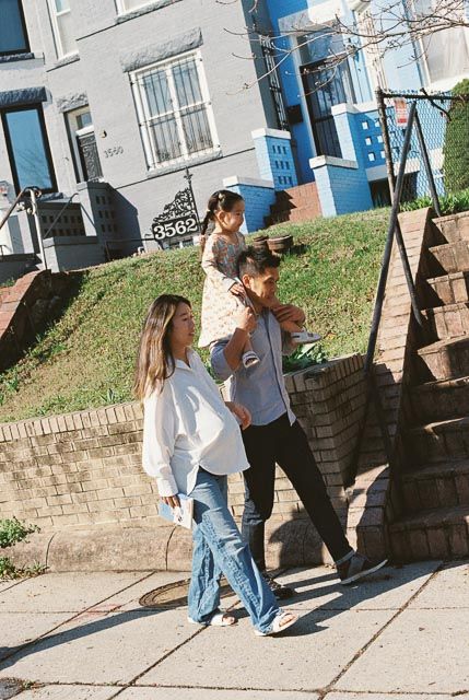 A family walks on a city sidewalk, with a child on a man's shoulders, beside brick retaining walls and colorful row houses.