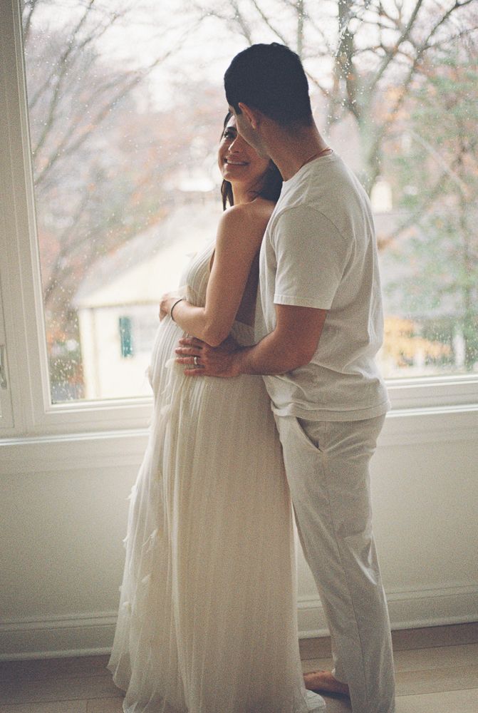 A romantic couple embraces by a bright window, with the person in white dress and other in casual white attire.