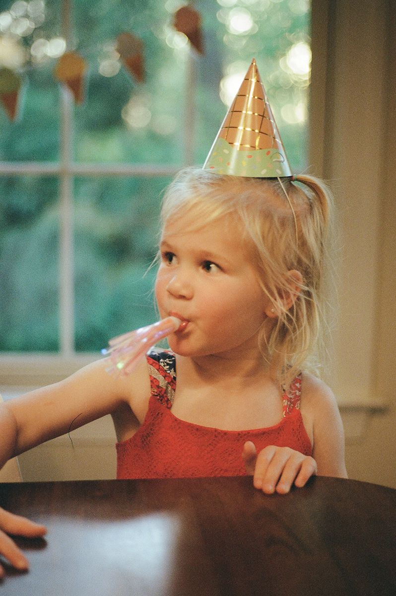A small child wearing a gold party hat and red dress sits at a table during a celebration.