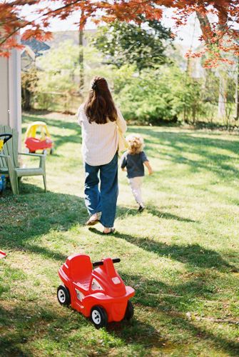 Mother and young child running together in a green backyard with a red ride-on toy car in the foreground.
