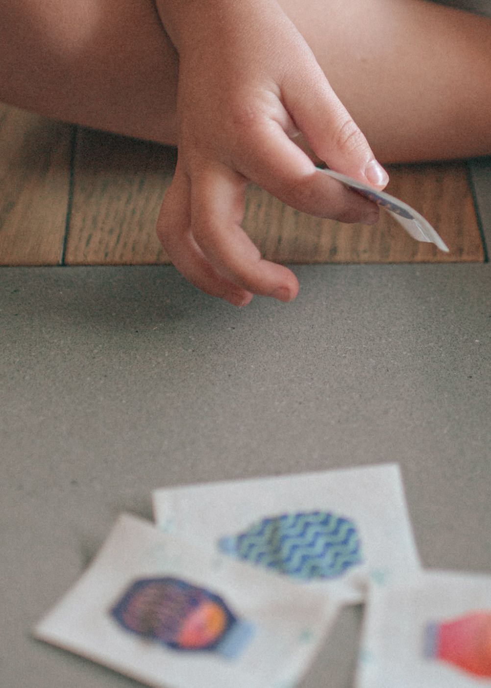 Hands carefully cutting paper crafts with colorful designs on a grey surface next to wooden flooring.