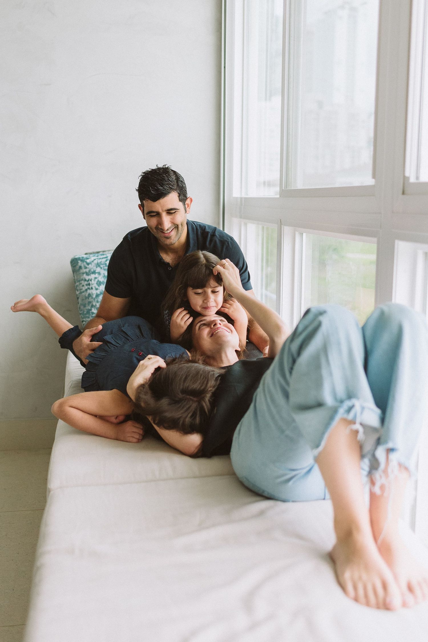 Family relaxing together on white bed by window with natural light streaming in.