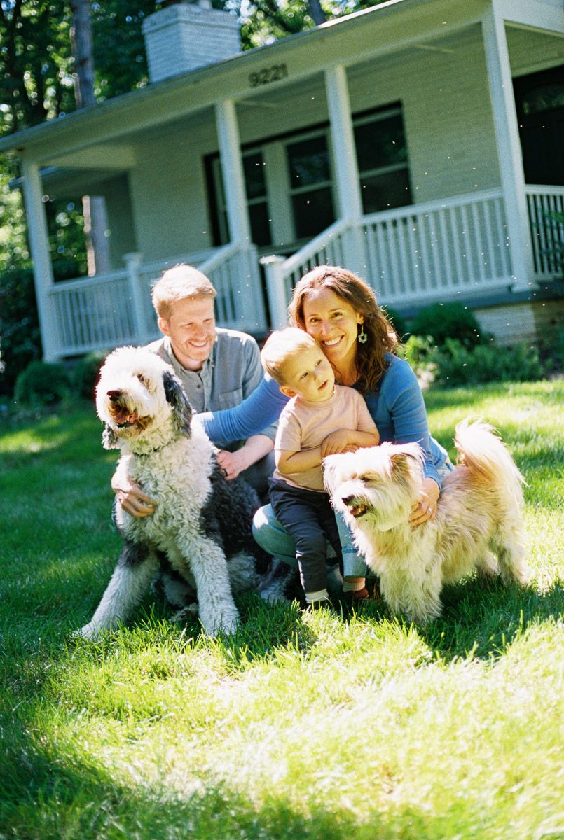 Happy family with two fluffy dogs posing on green lawn in front of charming suburban home on sunny day.