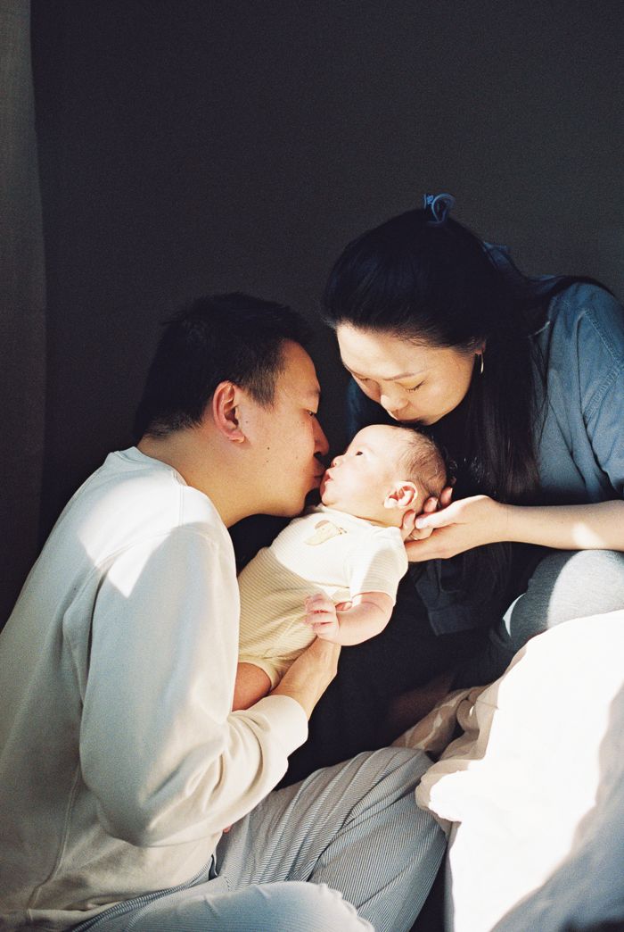 Two parents lovingly kiss and hold their newborn baby in a warm, intimate family portrait.