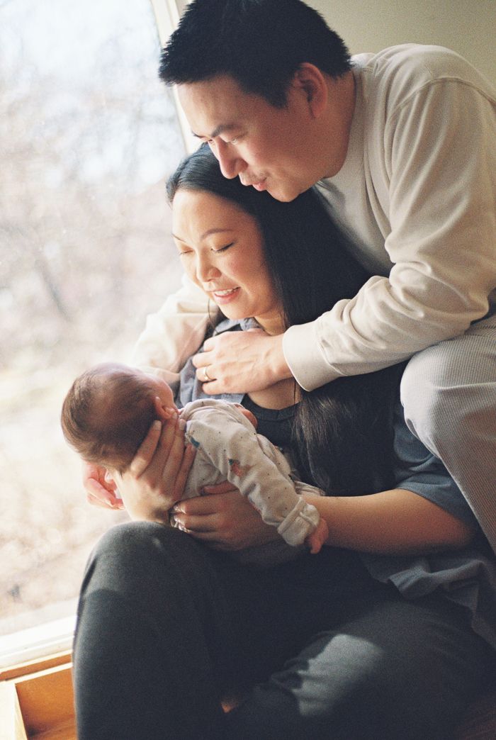 A couple lovingly gazes at their newborn baby by a sunlit window in a warm family portrait.