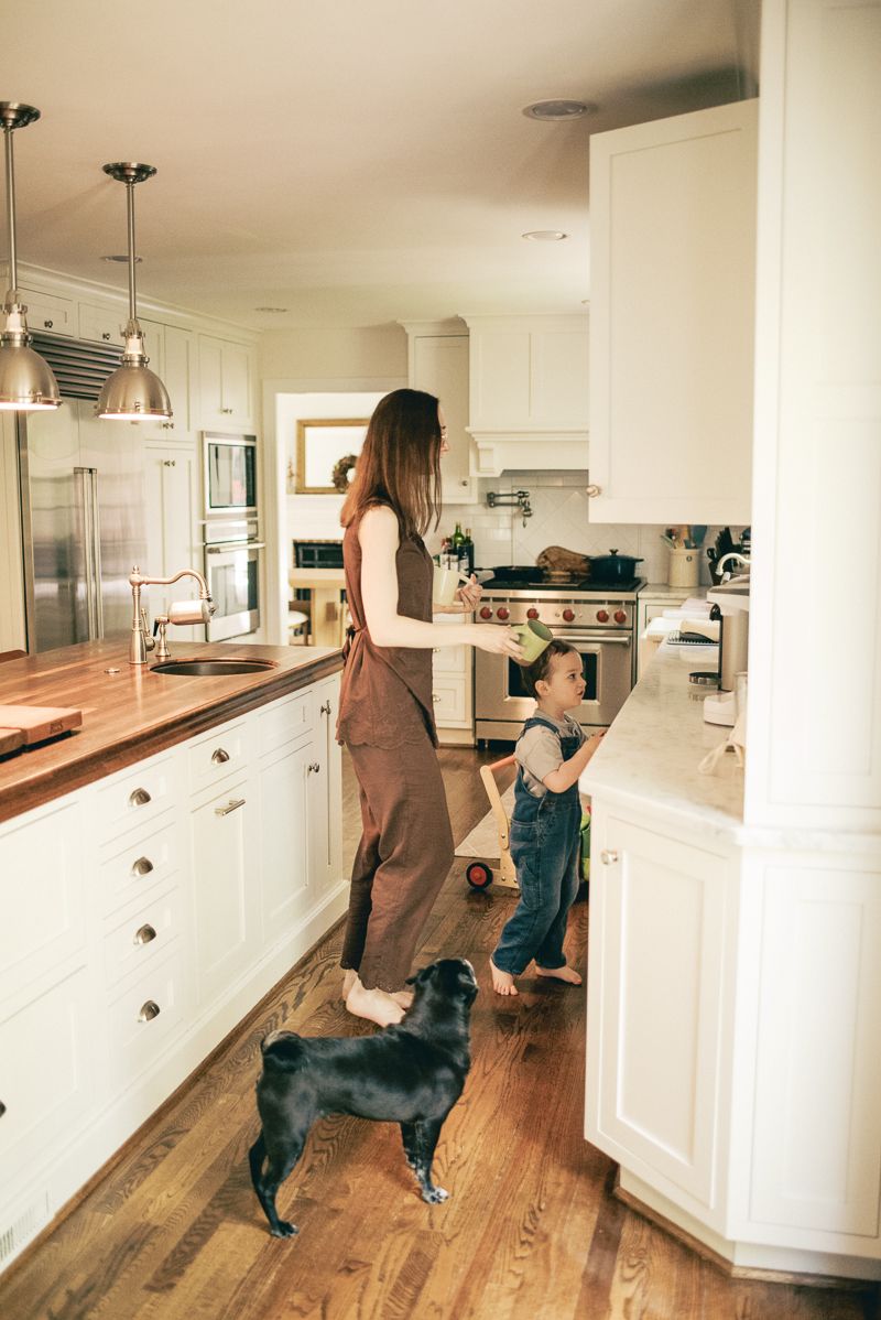 Mother in apron cooking with toddler and black dog in bright white farmhouse kitchen with wood floors.