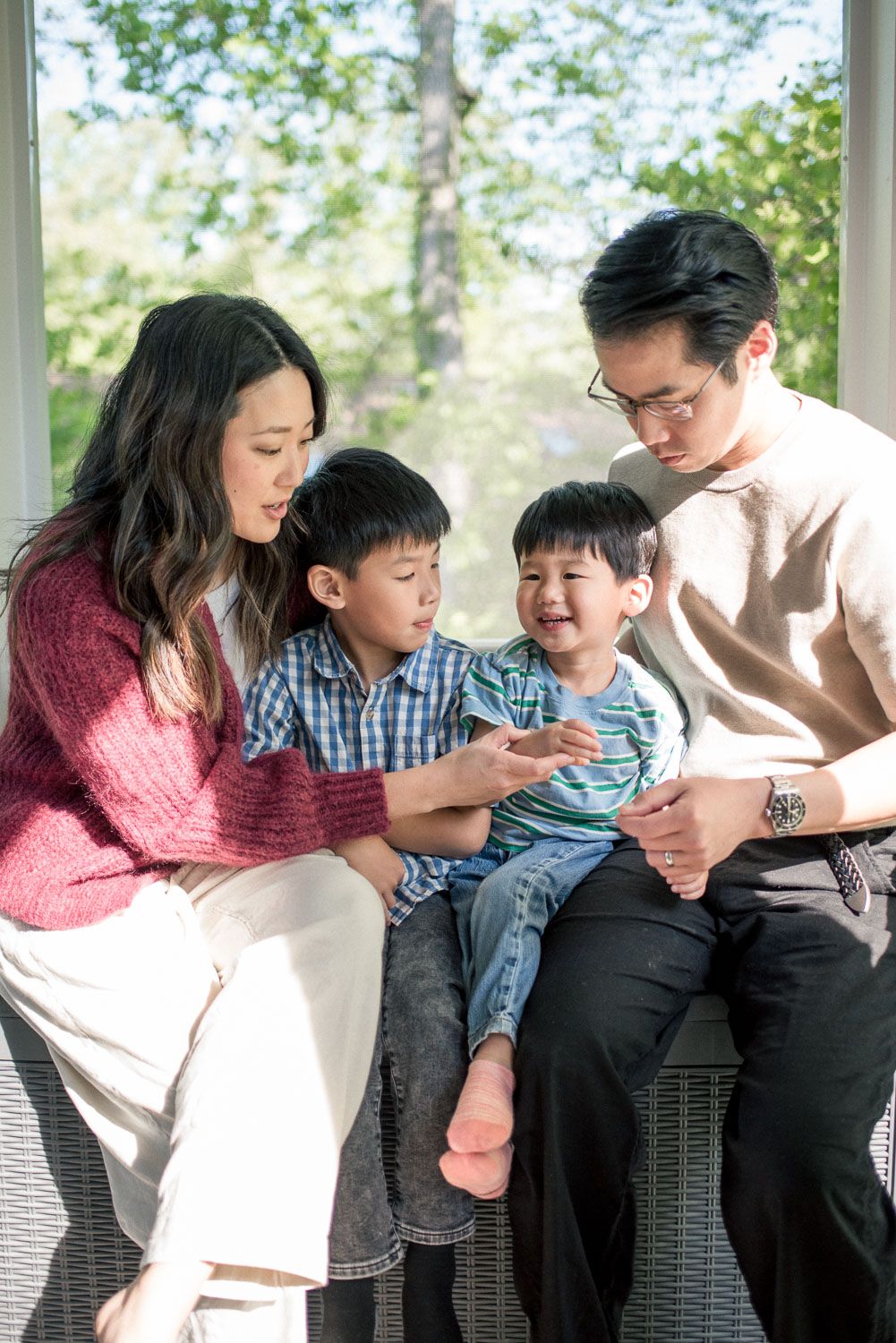 Asian family of four sitting together indoors, parents smiling at two young boys in a sunlit room with green trees outside.
