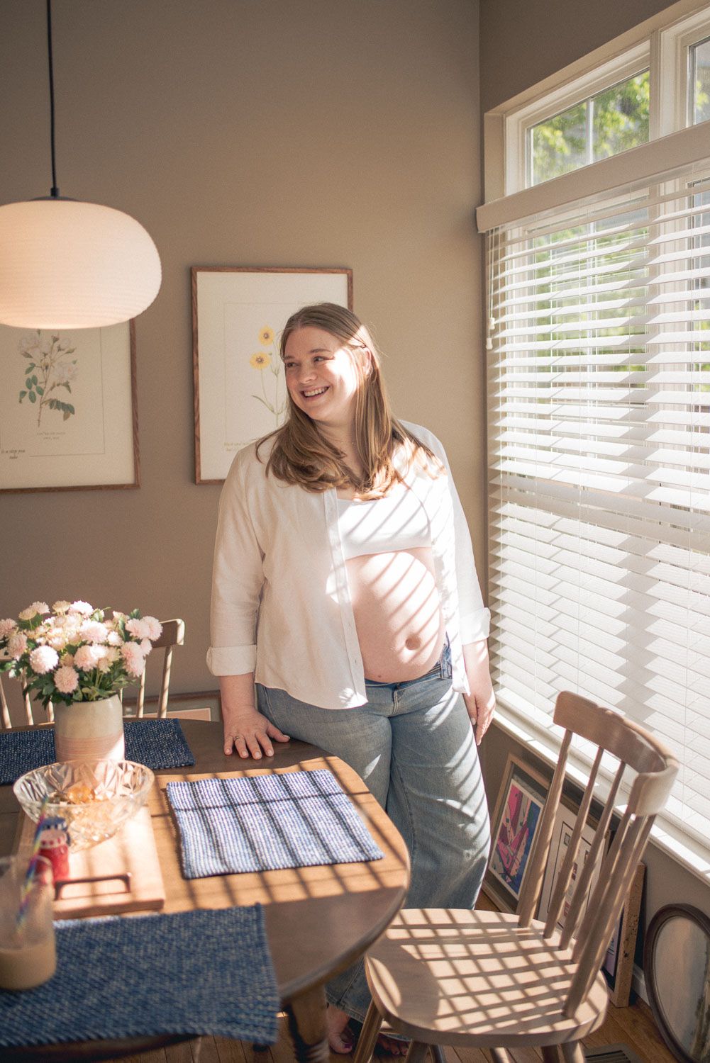 Smiling pregnant woman in white shirt and jeans stands by sunny dining room window, cradling her baby bump.