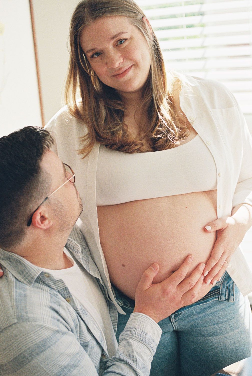 A smiling pregnant woman stands as her partner gently cradles her bare baby bump during a tender maternity shoot.