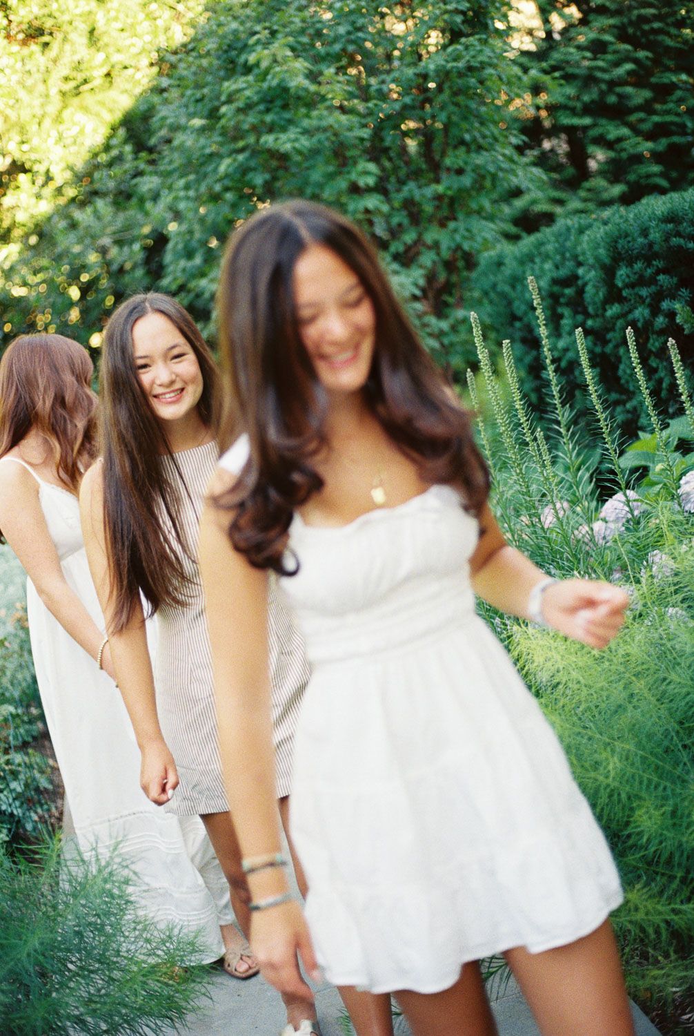 Three smiling women in white summer dresses walk single file along a garden path, surrounded by lush green foliage and flowers.