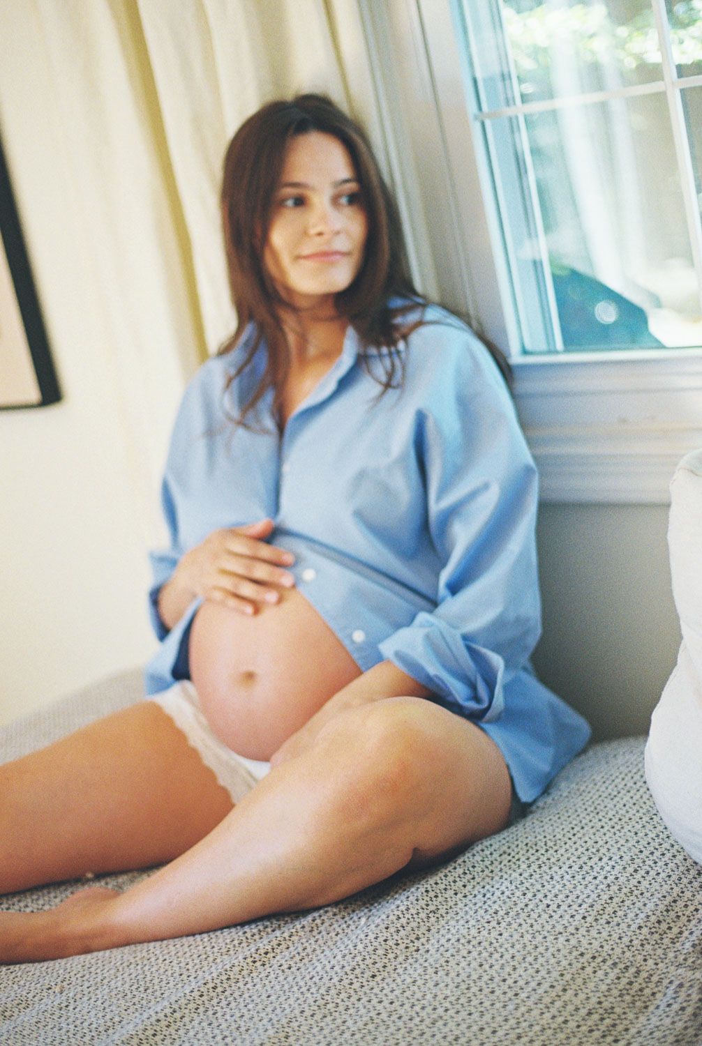 Pregnant woman in blue shirt sitting on bed cradling her baby bump near a sunlit window.