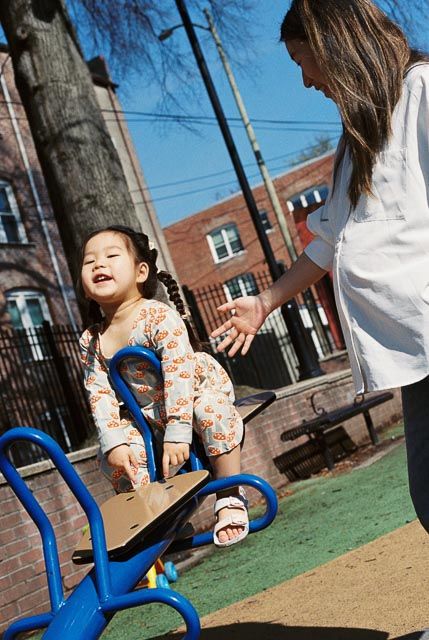A young girl laughs on a playground ride while a woman reaches out to her on a sunny day.
