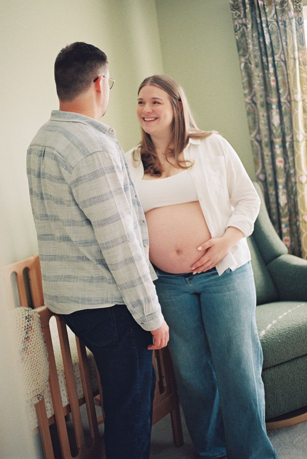 Smiling pregnant woman in white shirt and jeans stands with partner in cozy room with green sofa.