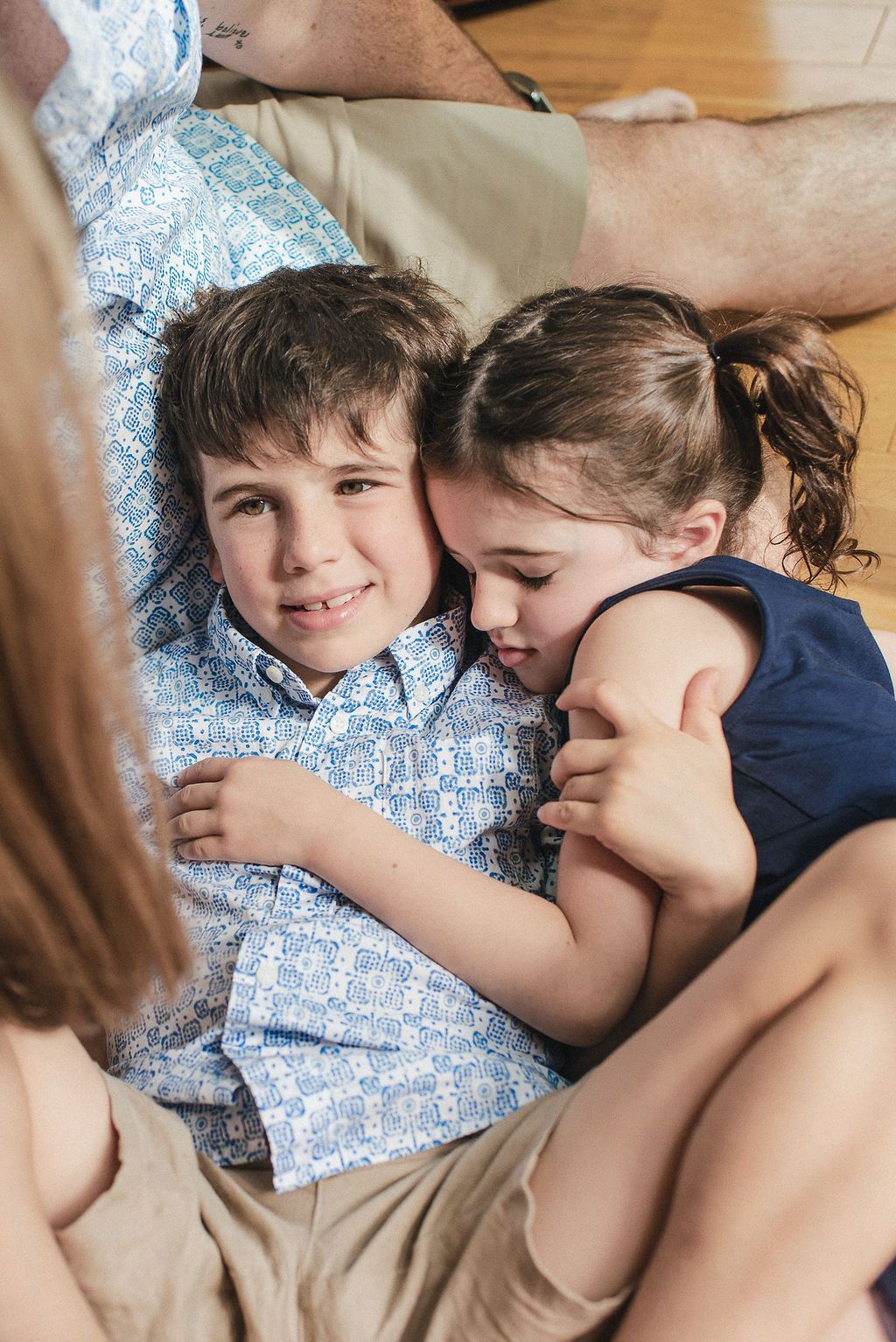 Two children cuddle together on a couch, one wearing a blue and white patterned shirt and the other in dark blue.