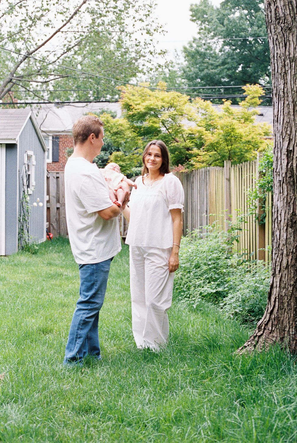 A couple stands in a lush backyard, man holding a newborn baby while woman smiles warmly near a large tree.
