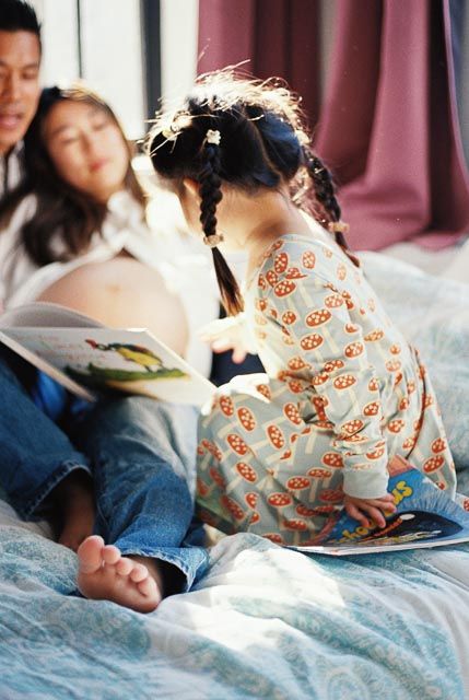 A young girl in mushroom print pajamas reads a book with her parents on a sunlit bed.
