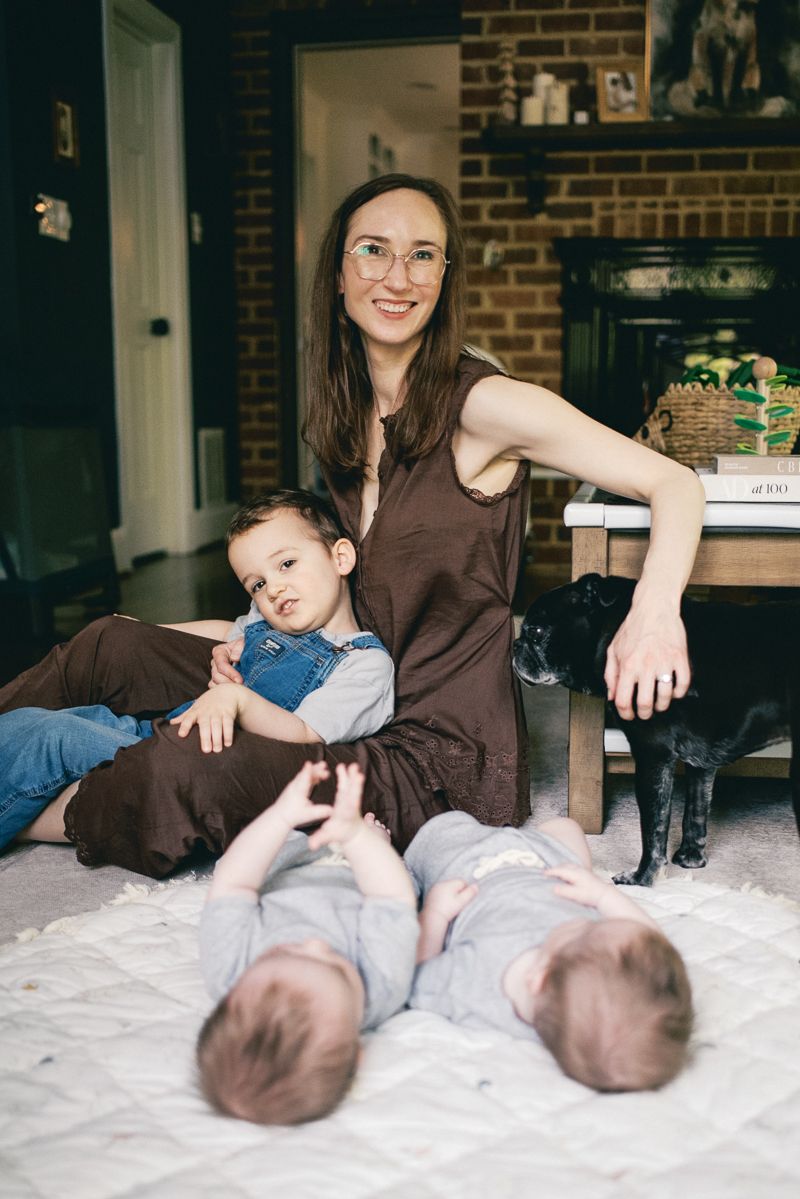 A smiling mother in glasses sits on the floor with a toddler and twin babies on a quilt, with a black dog nearby.