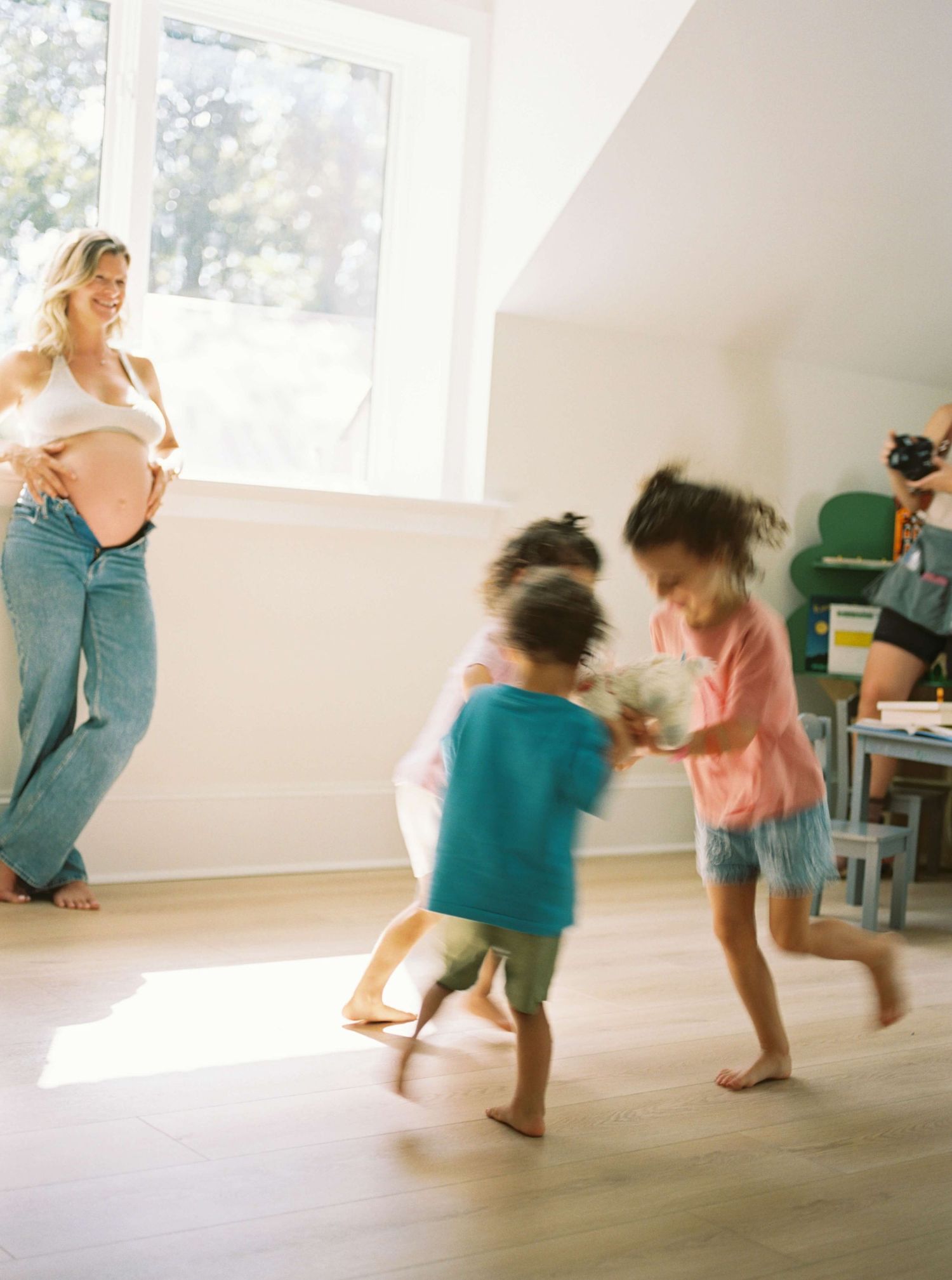 Children playing and dancing in sunlit room with large windows.