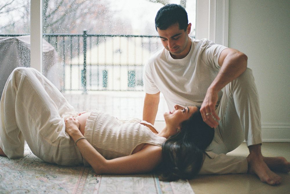 A couple relaxes together on a carpeted floor near a window in casual white clothing.