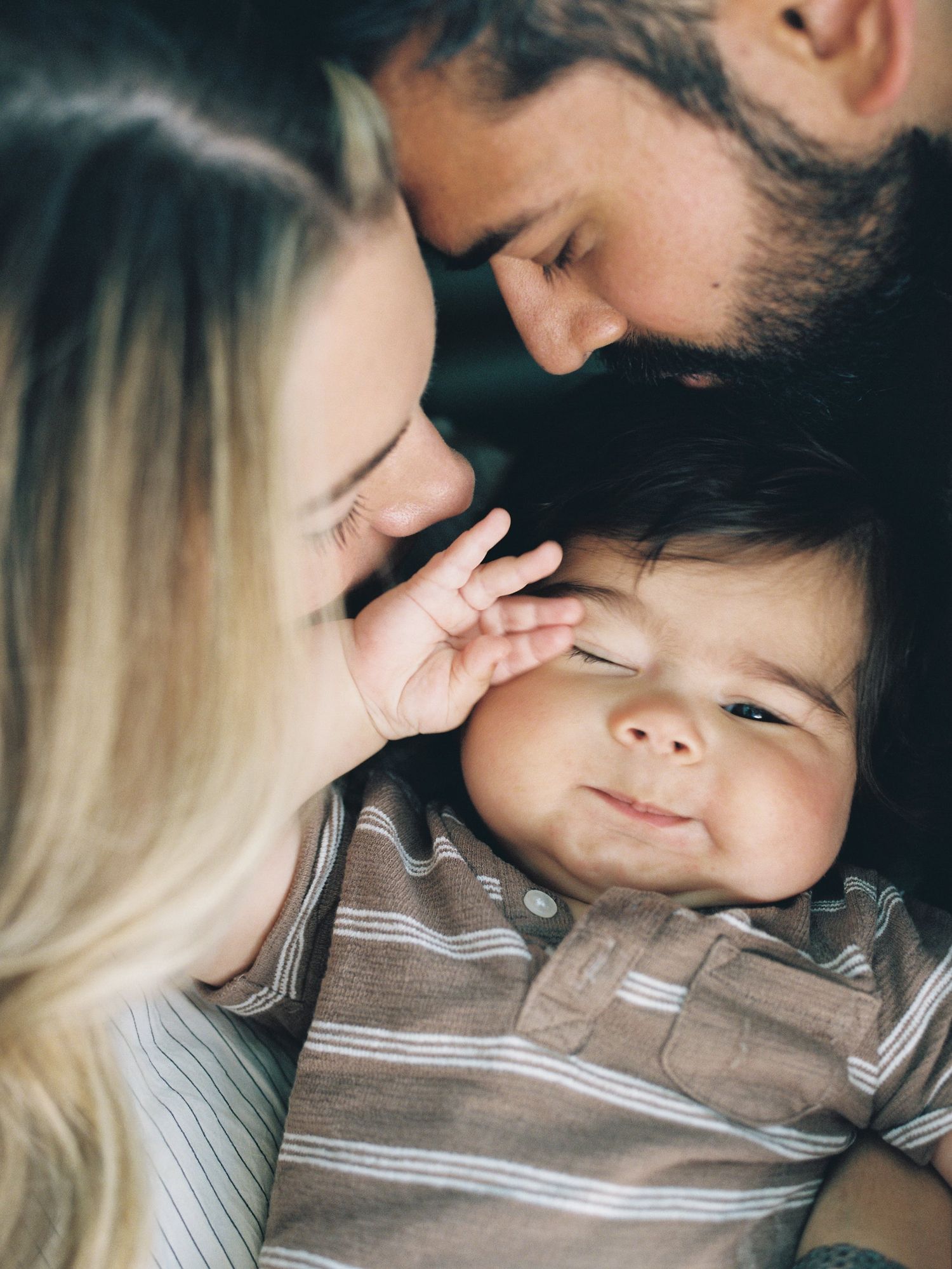 Intimate family portrait of parents sharing tender moment with smiling baby in striped shirt.
