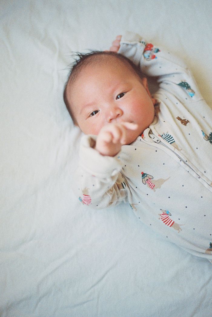 A newborn baby in a patterned onesie lies on white sheets, reaching one tiny hand toward the camera.