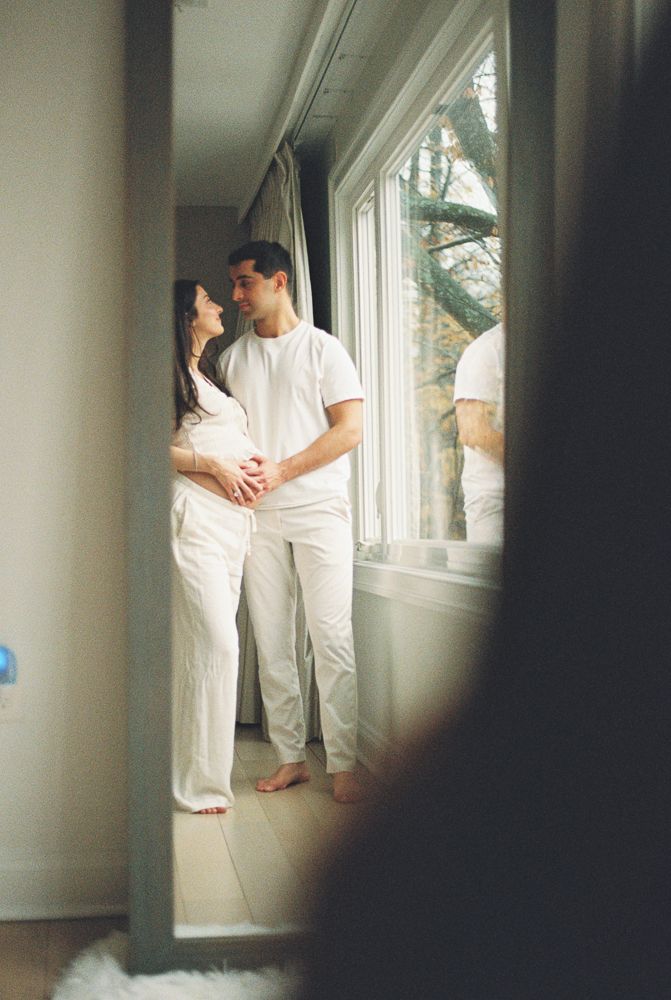 A couple in all white clothing stands intimately by a sunlit window in a minimalist interior space.
