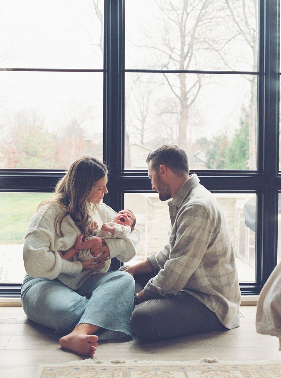 A couple sits cross-legged on the floor by large windows, gazing lovingly at their newborn baby.