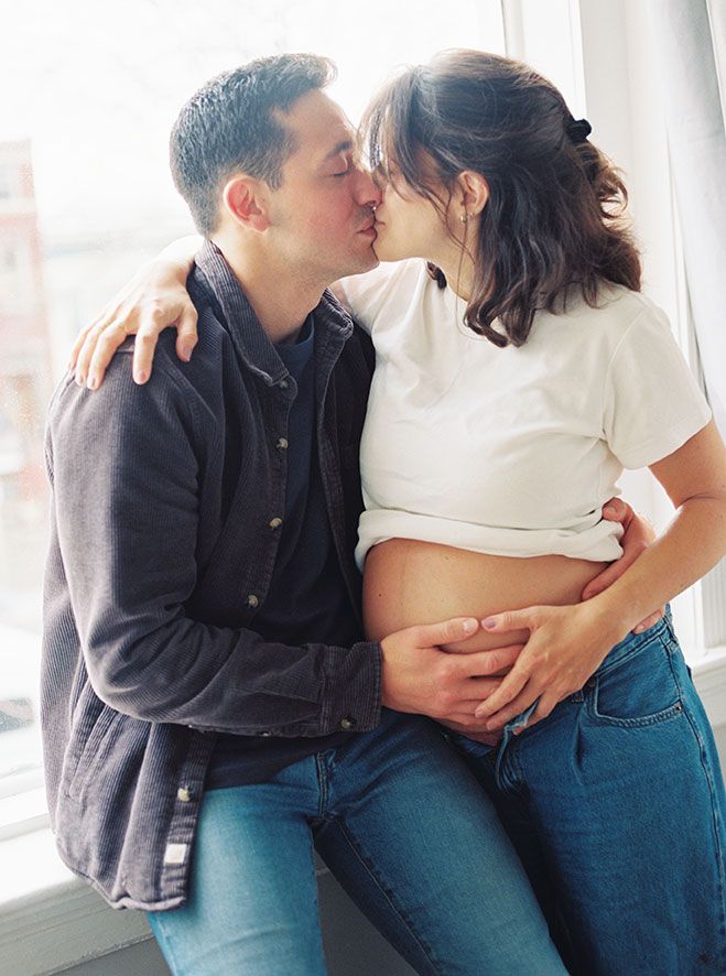 A couple in blue jeans and casual wear share an intimate moment by a bright window.