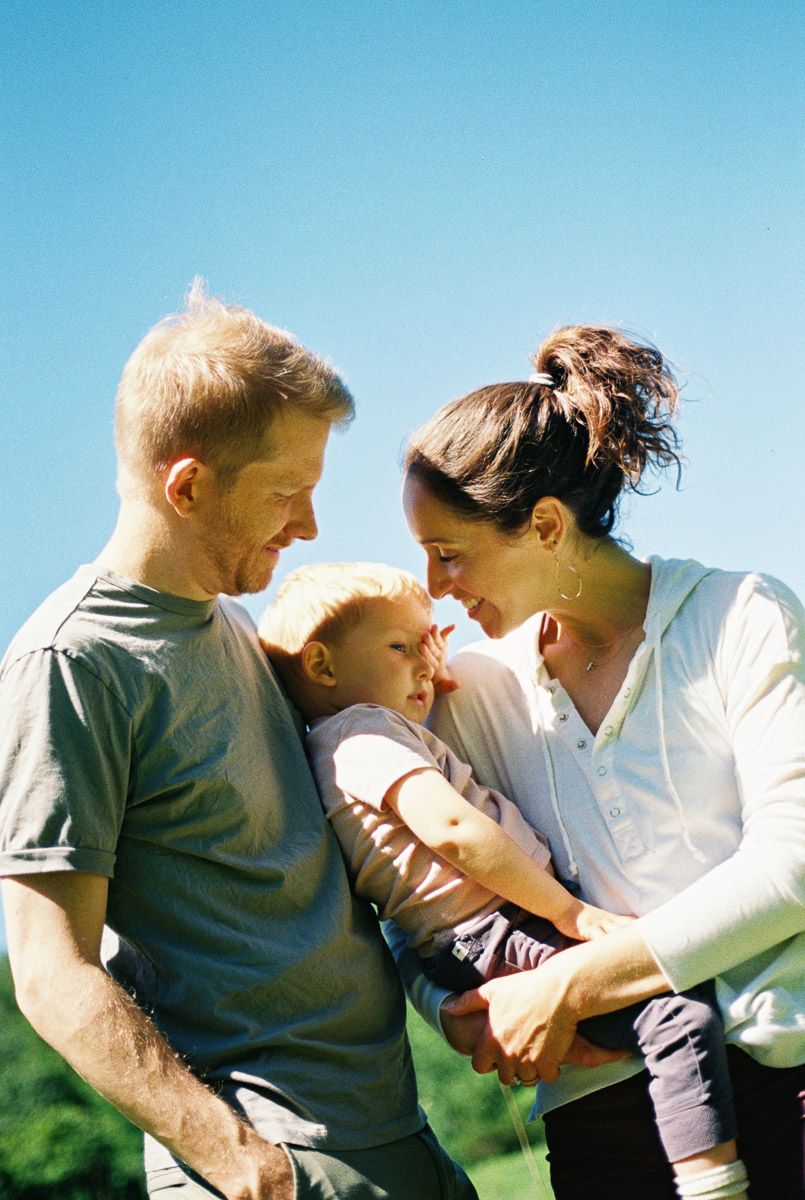 A happy young family with a baby enjoys a sunny outdoor moment, parents leaning in close together with smiles.