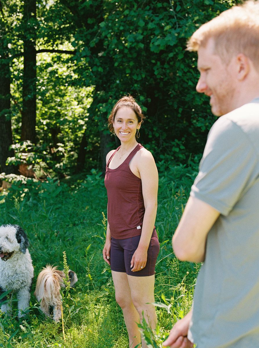 A smiling woman in a maroon tank top and shorts stands in a lush green forest with two dogs beside her.