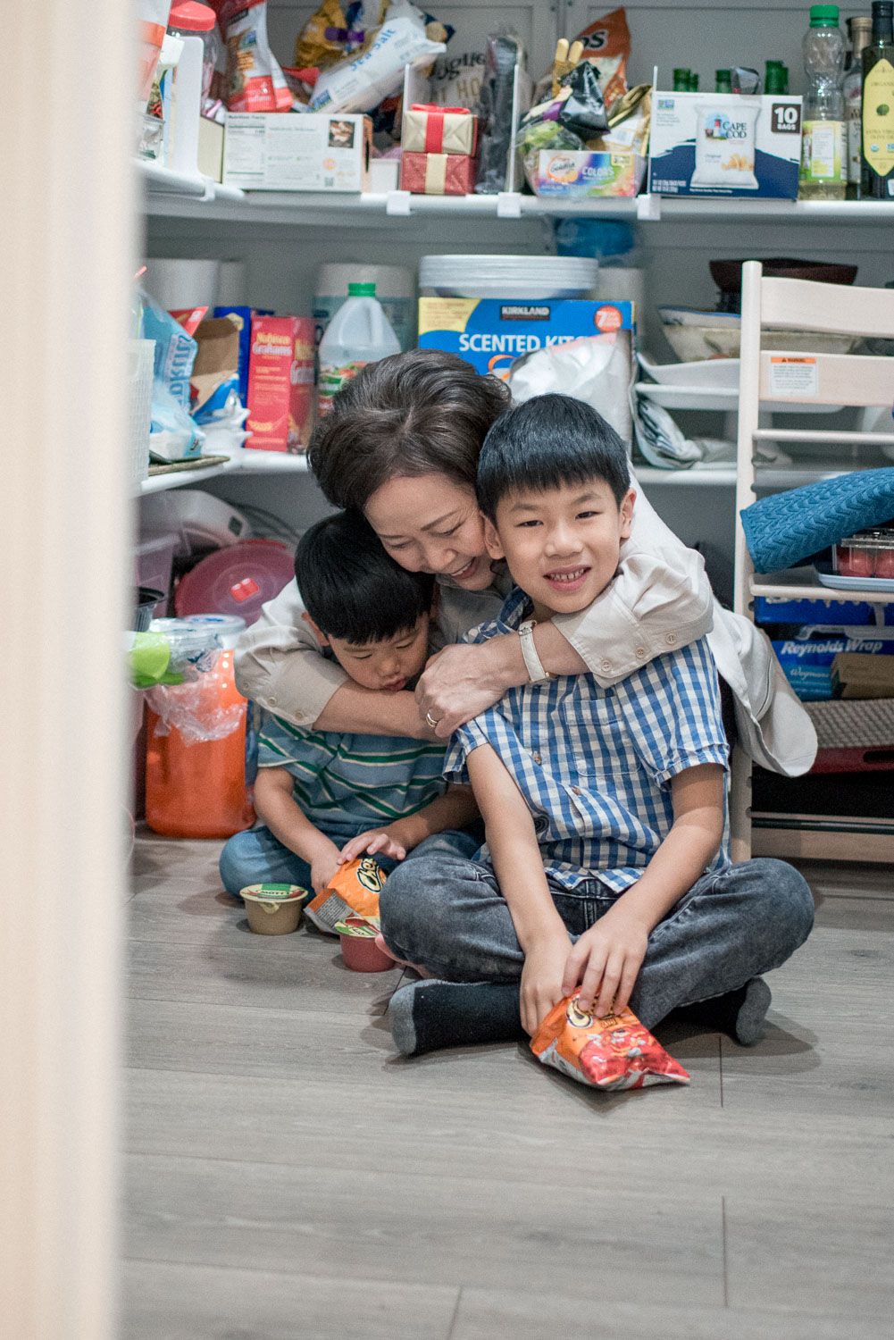 Asian mother hugs two young sons on pantry floor, sharing a joyful family moment with snacks.