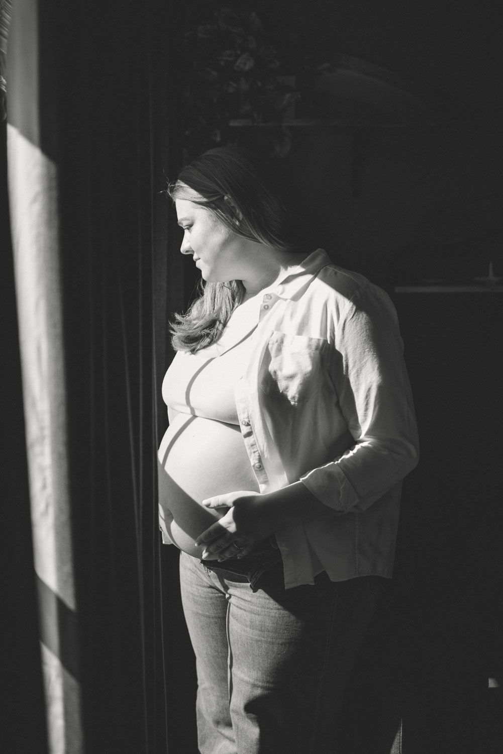 Black and white photo of a pregnant woman cradling her baby bump while standing by a window in soft natural light.
