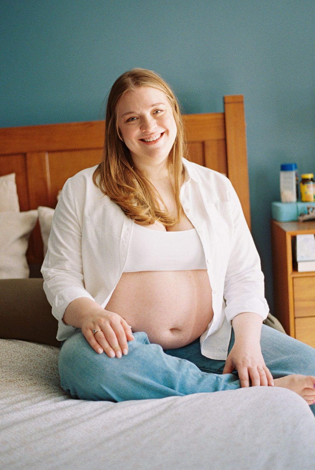 Smiling pregnant woman sitting on bed with exposed baby bump, wearing white shirt and jeans.