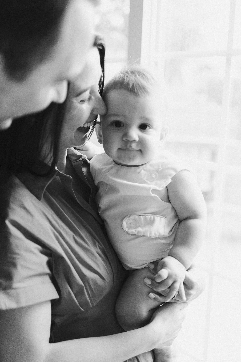 Tender black and white portrait sequence of a parent holding their baby by a bright window.