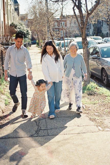 Asian family of four walking on sunny sidewalk, grandma and parents holding toddler girl's hands in residential neighborhood.