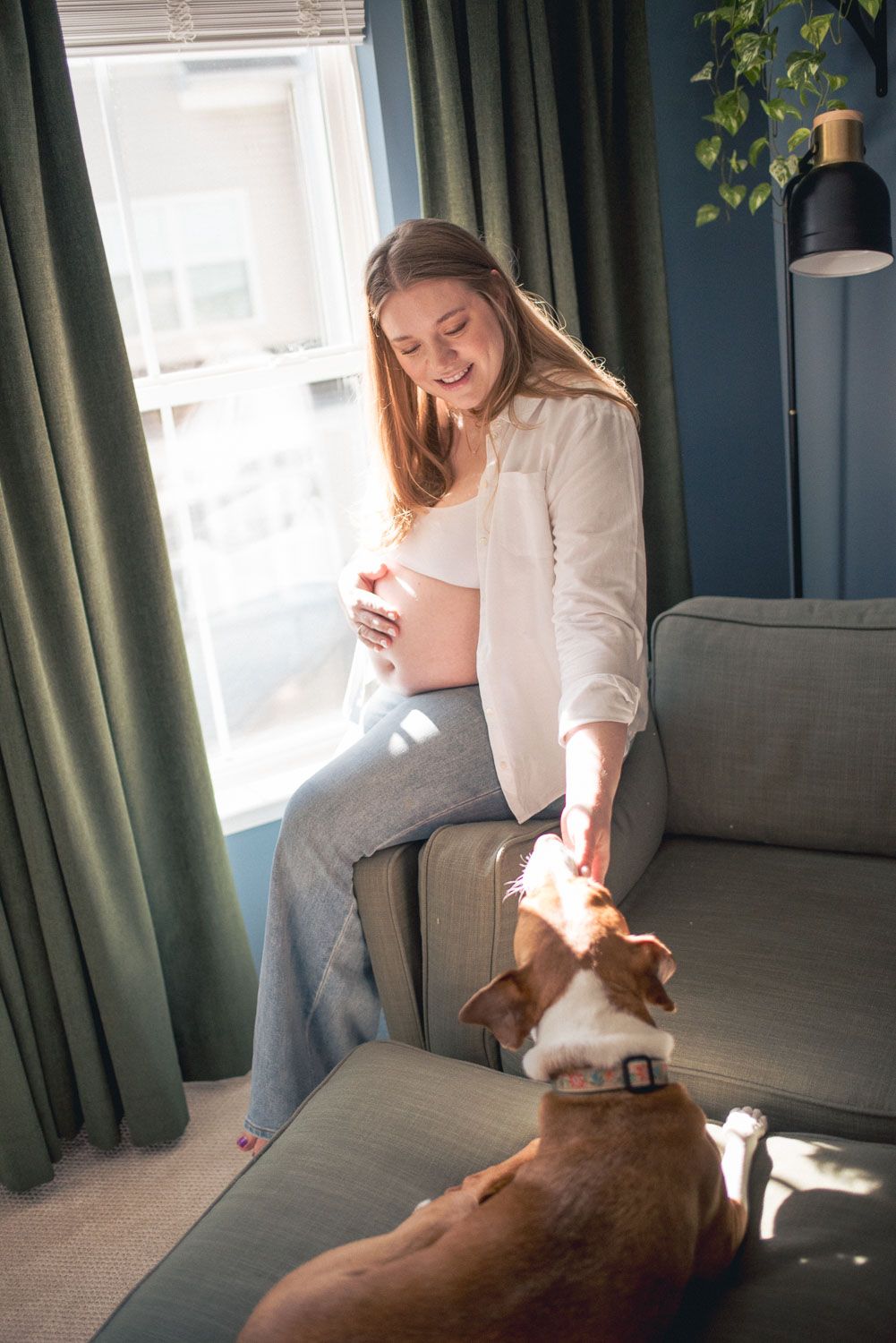 Smiling pregnant woman in white shirt sits on sofa petting a brown and white dog by a sunlit window.