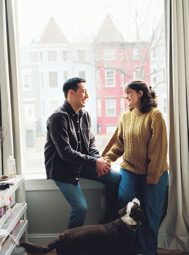 A couple sits and chats by a bright window in a cozy indoor setting wearing casual winter attire.