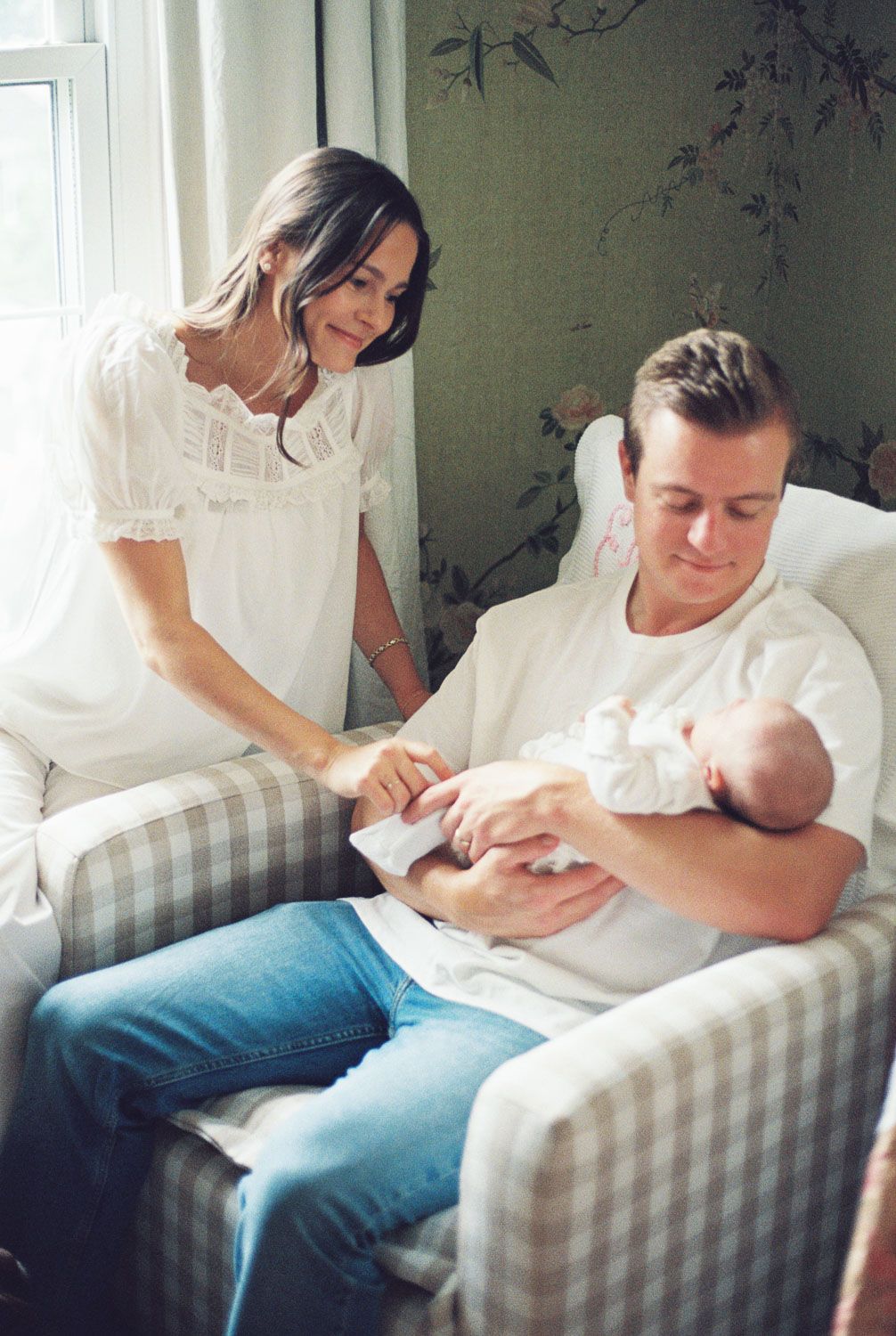 A couple in white outfits admires their newborn baby in a cozy nursery chair by a sunlit window.
