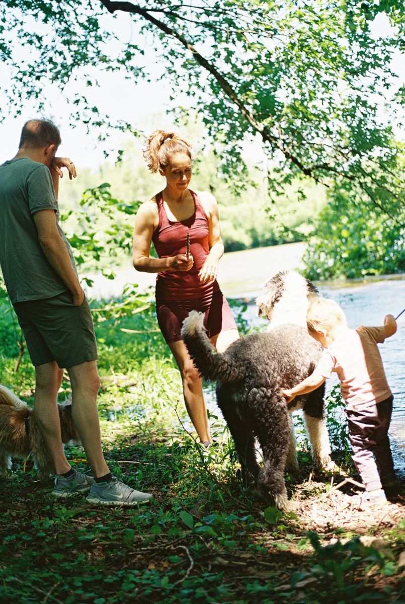 Woman in red swimsuit with a gray poodle near a river, surrounded by lush green trees on a sunny day.