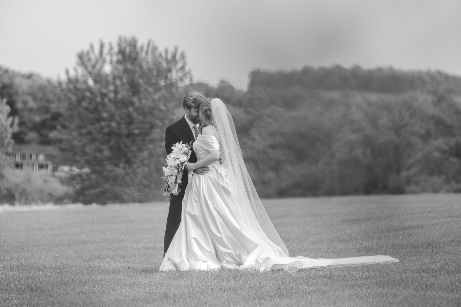 Black and white photo of a bride and groom embracing on a green lawn with trees in the background.