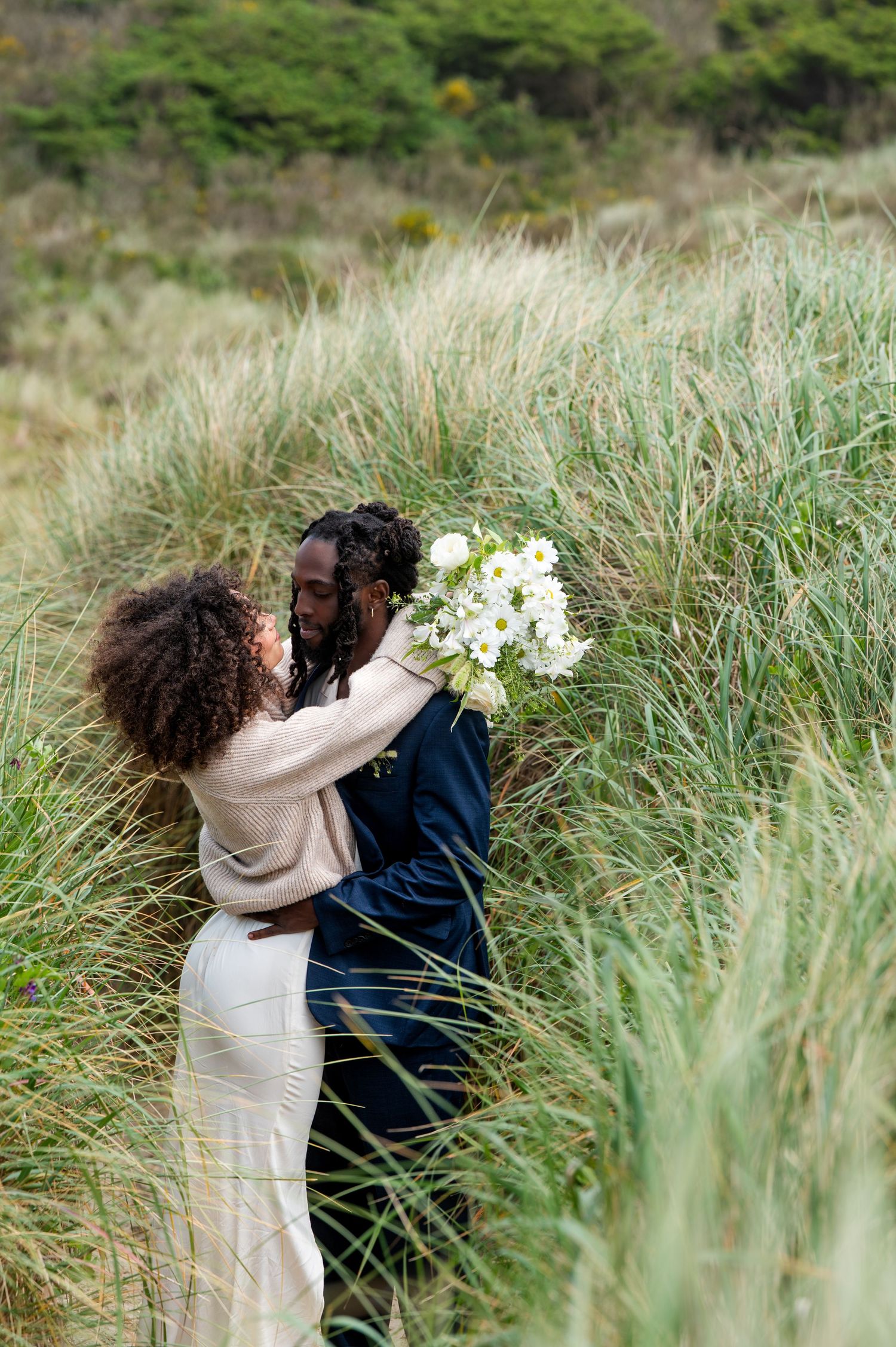 A couple embraces in tall grass, the bride holding a white bouquet, sharing a romantic moment outdoors.