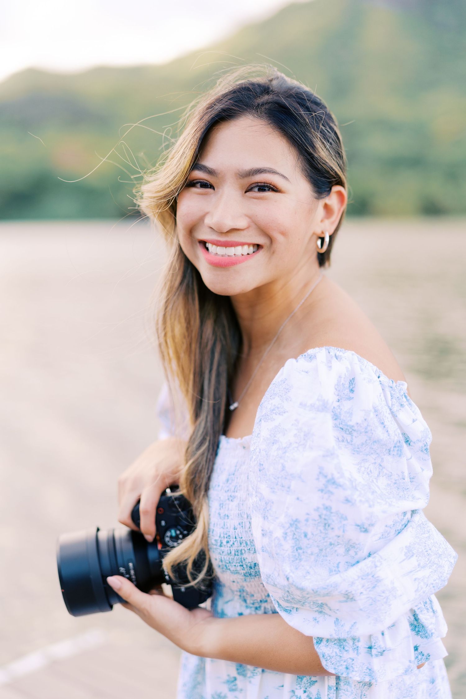 A photographer in a delicate white and blue floral dress holds a professional camera while standing outdoors.