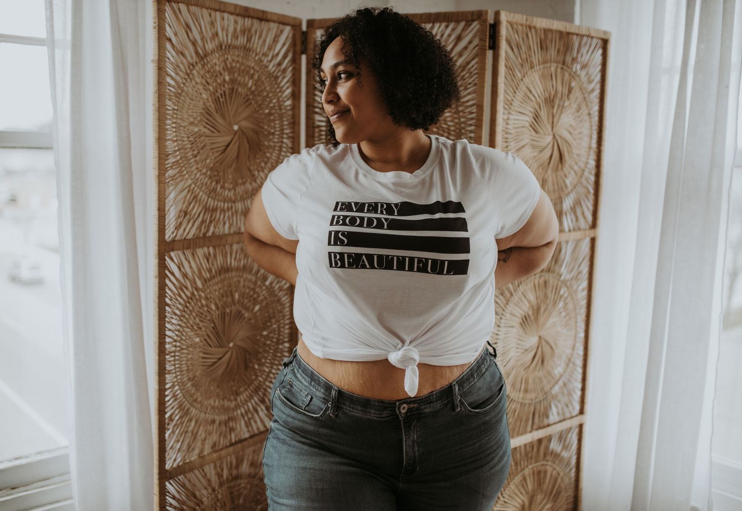 Woman in white crop top and gray pants stands against textured wall in natural light setting.