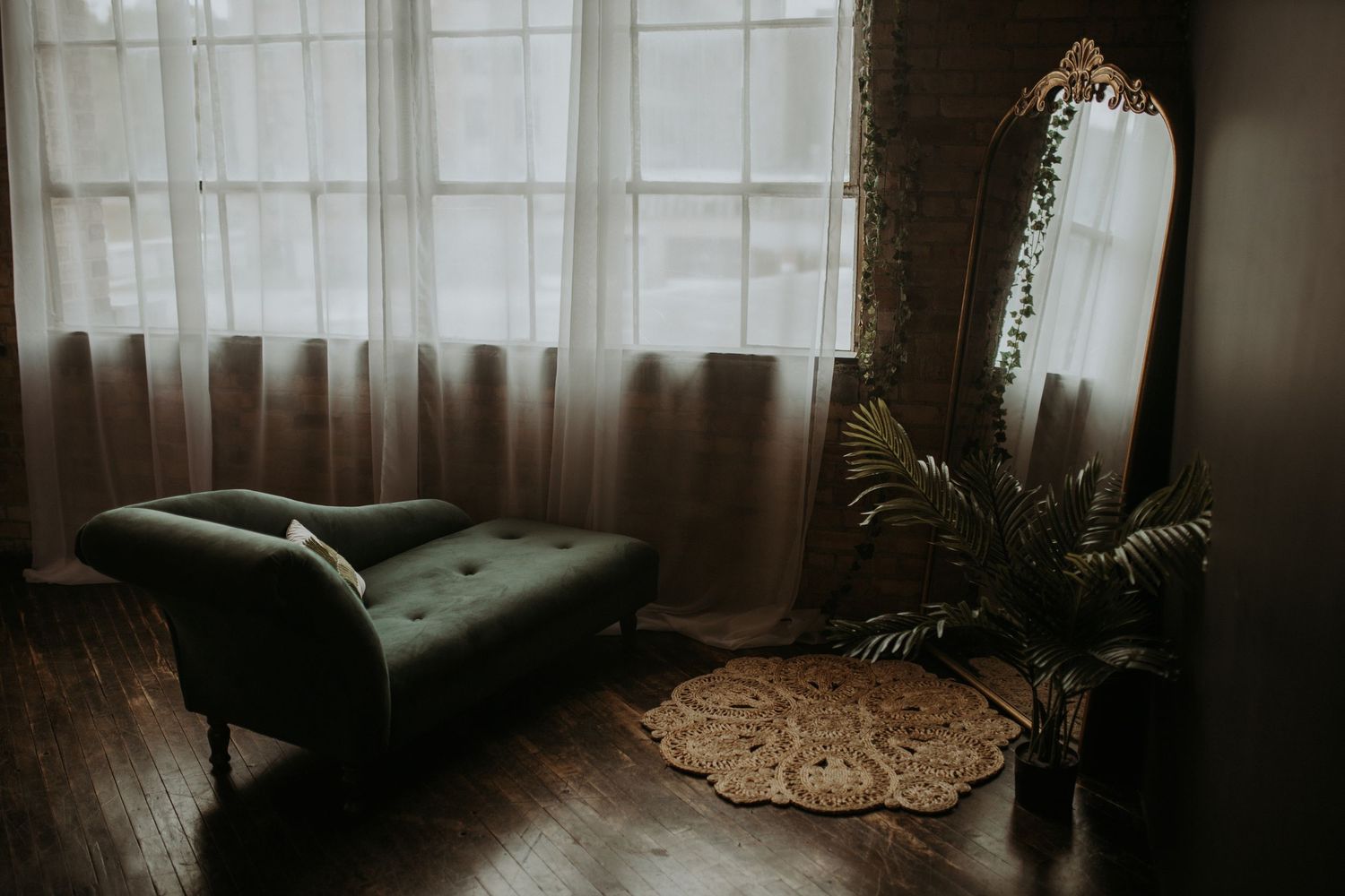 Moody interior scene with vintage green armchair, tall mirror, potted plant and jute rug on dark hardwood floors.
